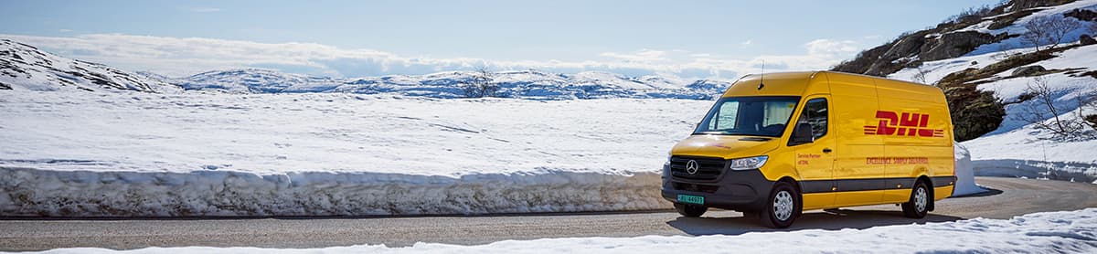 Furgoneta amarilla de DHL circulando por una carretera nevada, rodeada de un paisaje invernal.